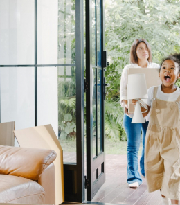 Happy African American young family bought new house. Mom, Dad, and child smiling happy hold cardboard boxes for move object walking into big modern home. New real estate dwelling, loan and mortgage.
